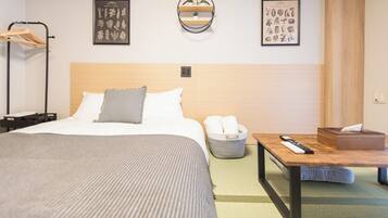 A hotel room with a bed, a bedside table with a remote control, a basket of towels, and two framed pictures on the wall.