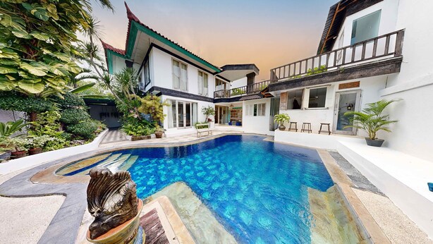 A swimming pool with a stone sculpture, surrounded by lush greenery and a two-story building with a balcony. - Villa Kendal Seminyak (Seminyak)