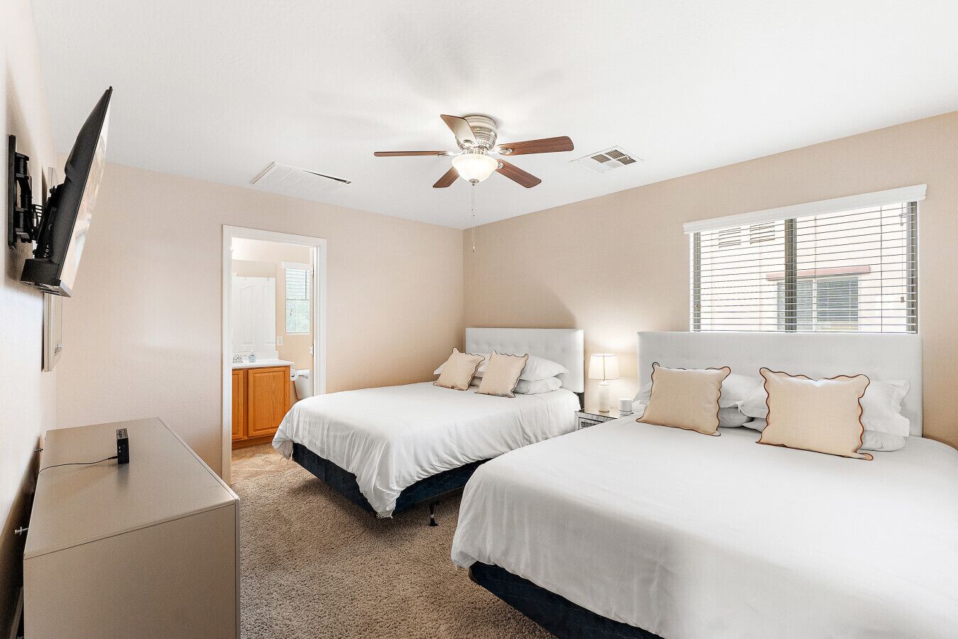 A hotel room with two beds, a ceiling fan, a TV mounted on the wall, and a window with blinds.