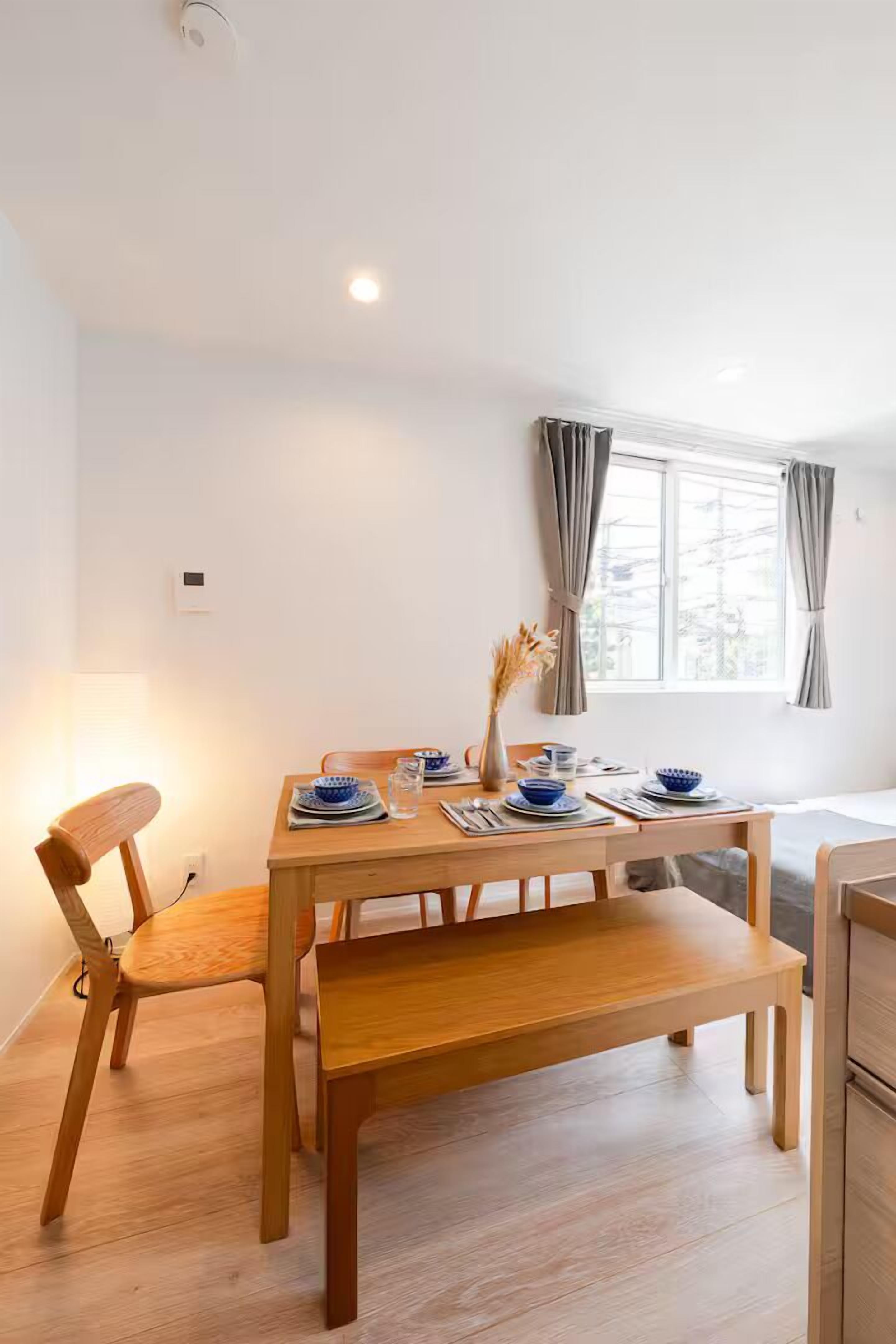 Een eetruimte met een houten tafel gedekt voor twee personen, een bankje en een raam met gordijnen.