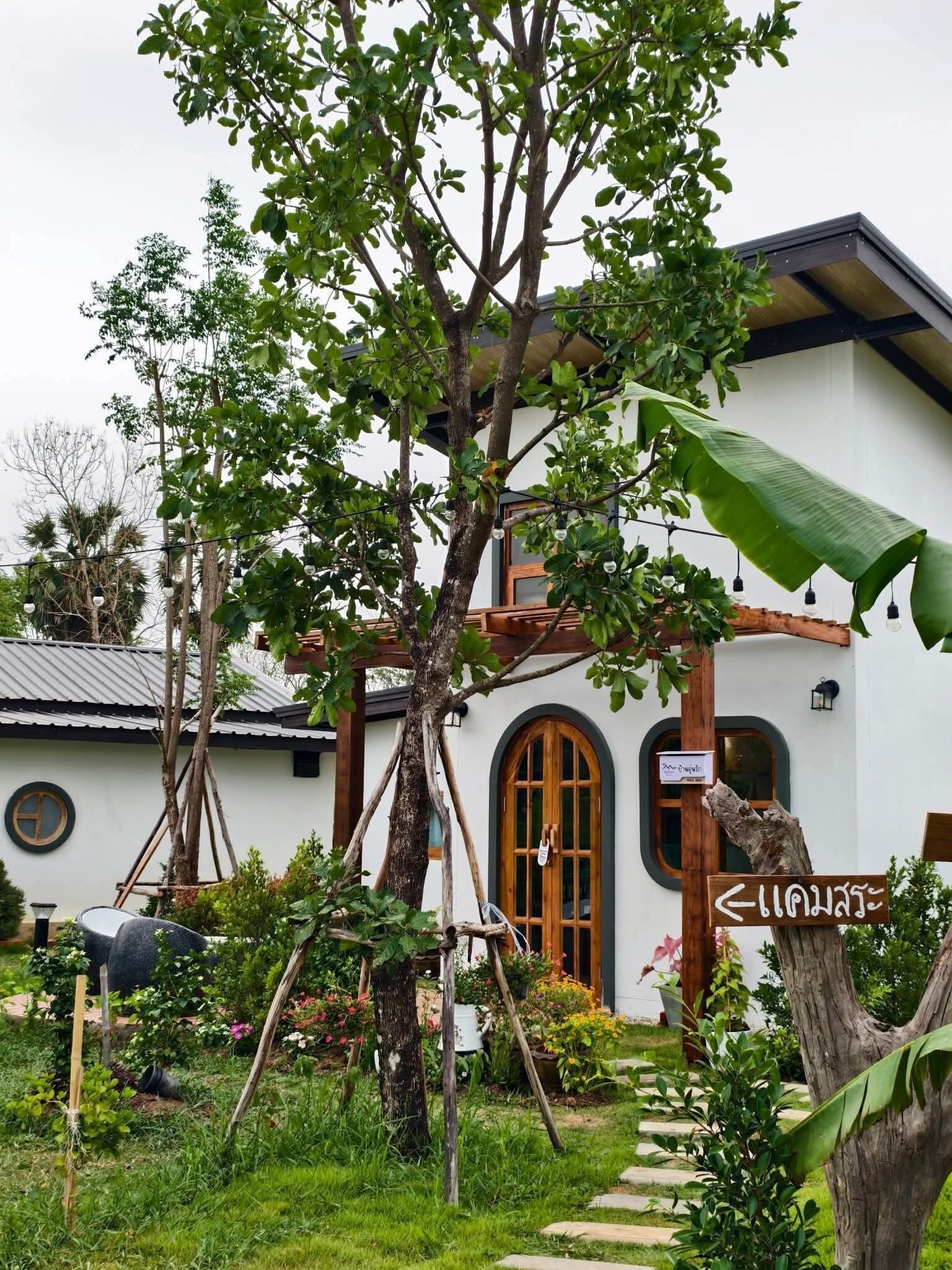 A white building with a wooden door and windows, surrounded by lush greenery and a garden.