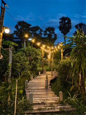 Un sendero de madera serpentea por un jardín exuberante al anochecer, con luces colgantes.