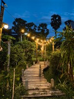 Un sendero de madera serpentea por un jardín exuberante al anochecer, con luces colgantes.