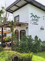 Un edificio blanco con un logotipo verde y texto tailandés, una pérgola de madera con plantas trepadoras y una maceta con forma de rueda de bicicleta.