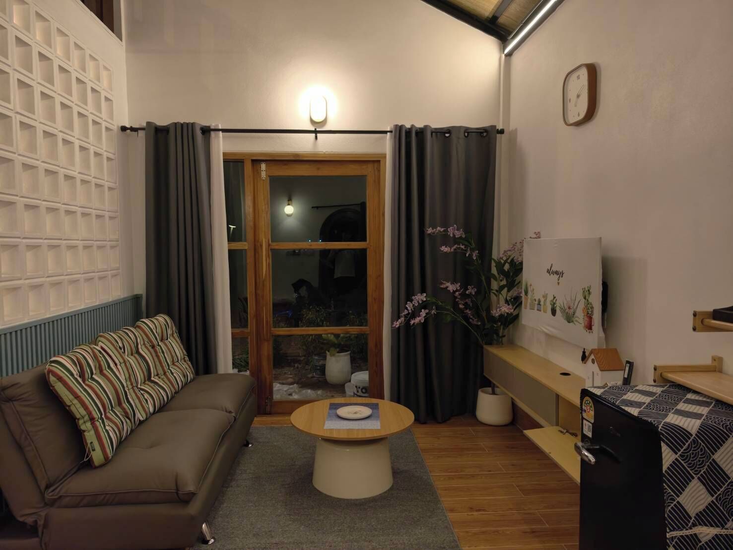 A living room with a sofa, a coffee table, a refrigerator, a clock, and a window with curtains.