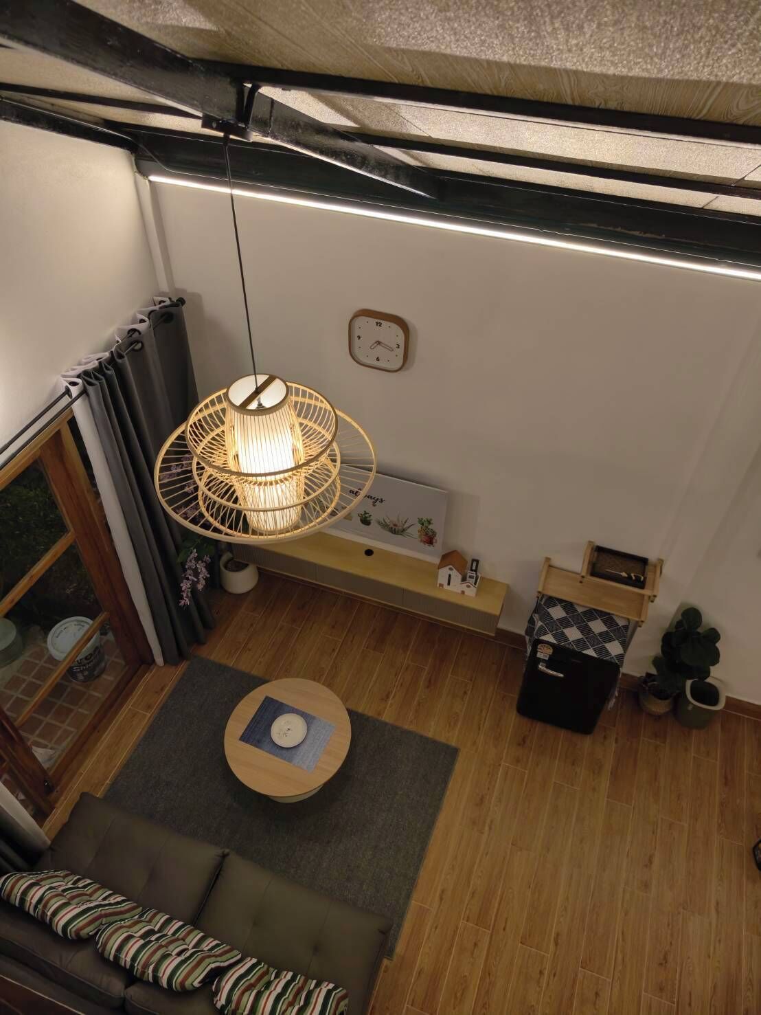 A compact living space with a sofa, a small round table, a hanging light fixture, a clock, and a shelf with decorative items.