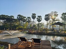 Terraza de madera con dos sillones de descanso y una mesita, con vista a un cuerpo de agua, un pabellón y palmeras al fondo.