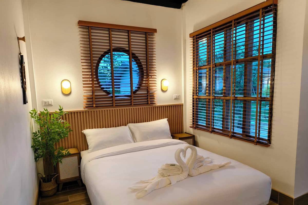 A hotel room with a bed, wooden headboard, two bedside tables, a potted plant, and two windows with blinds.