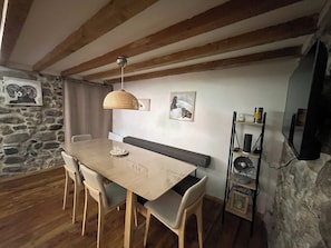 Un coin repas comprenant une table en bois, quatre chaises, un luminaire suspendu en fibres tressées, un banc et un mur en pierre.