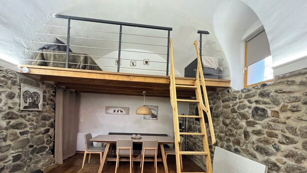 A loft-style room with a dining area, a wooden ladder, stone walls, and a view of the sky.