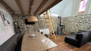 A spacious room with a wooden dining table, stone walls, a black leather sofa, and a wooden ladder.