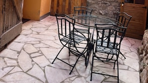 A glass-top table with metal chairs in a courtyard with stone flooring, surrounded by a yellow wall and a stone building.