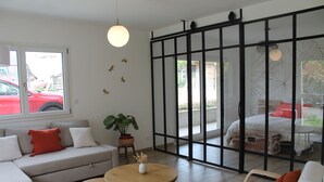 A modern living room with a sofa, two round tables, a potted plant, and a sliding glass door leading to a bedroom.