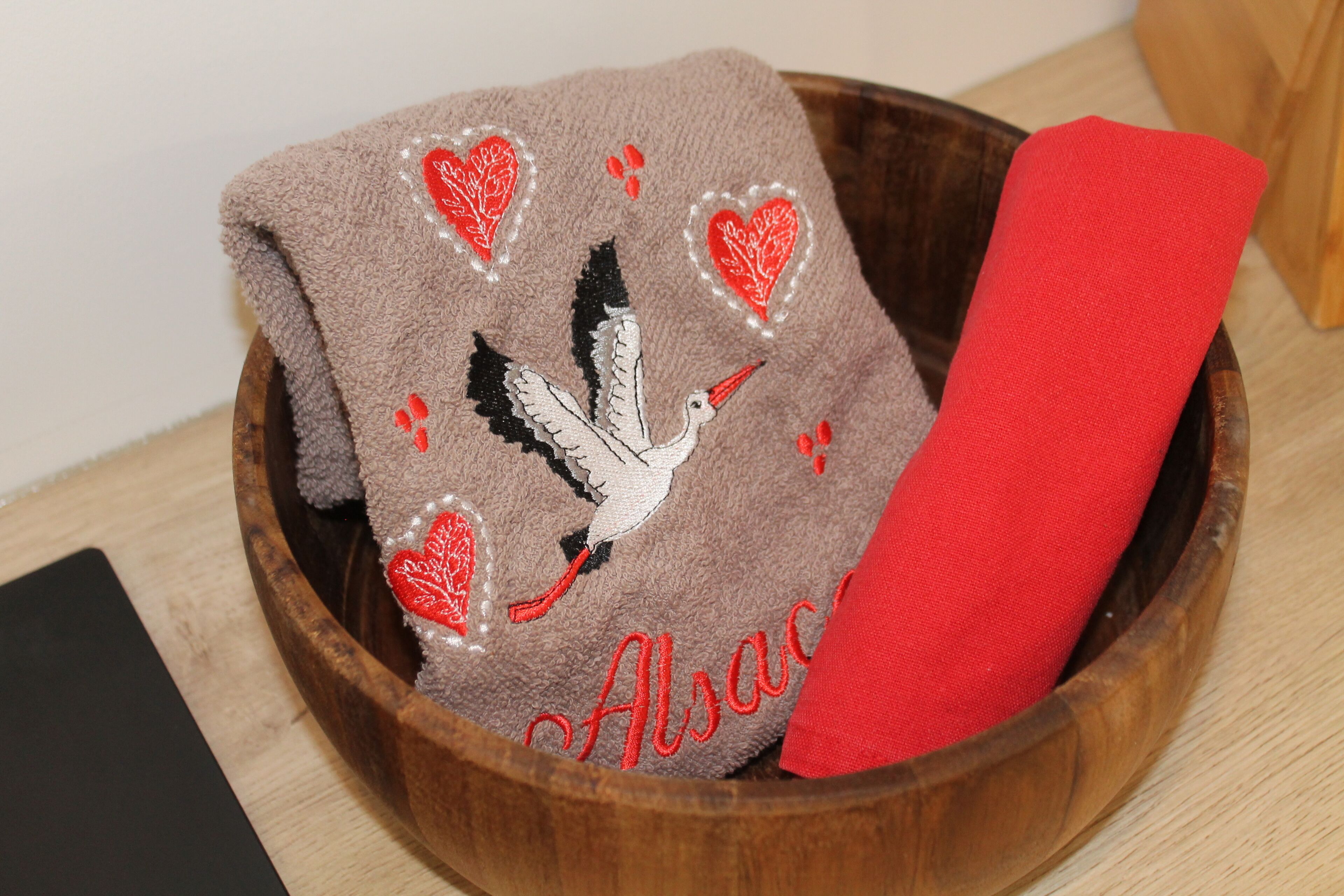 A wooden bowl containing a brown towel with embroidered birds and hearts, and a red cloth.