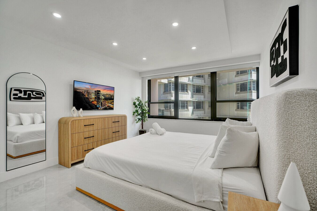 A modern bedroom with a large bed, a wooden wardrobe, a cityscape painting, and a large window with a view of buildings.
