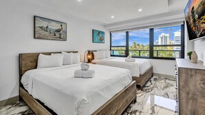 A hotel room with two beds, a large window with a city view, a painting on the wall, and a flat-screen TV. - Luxury Beachside 3BR/3BA Condo + Resort Amenities, Pools, Gym and Sauna 620 (Miami Beach)