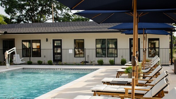 Un espace aménagé au bord de la piscine, avec des chaises longues et des parasols, une piscine, et un bâtiment doté de fenêtres, avec une clôture en arrière-plan.
