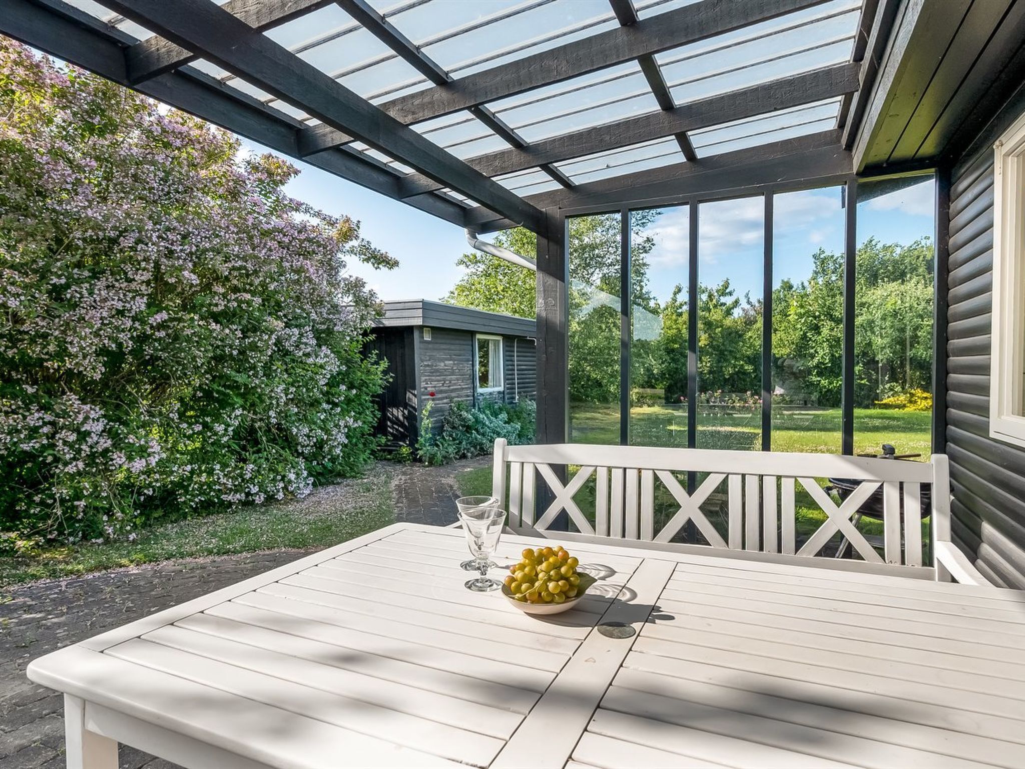 Ein überdachter Außenbereich mit Tisch und Bank, umgeben von Pflanzen und Blick auf einen Garten.