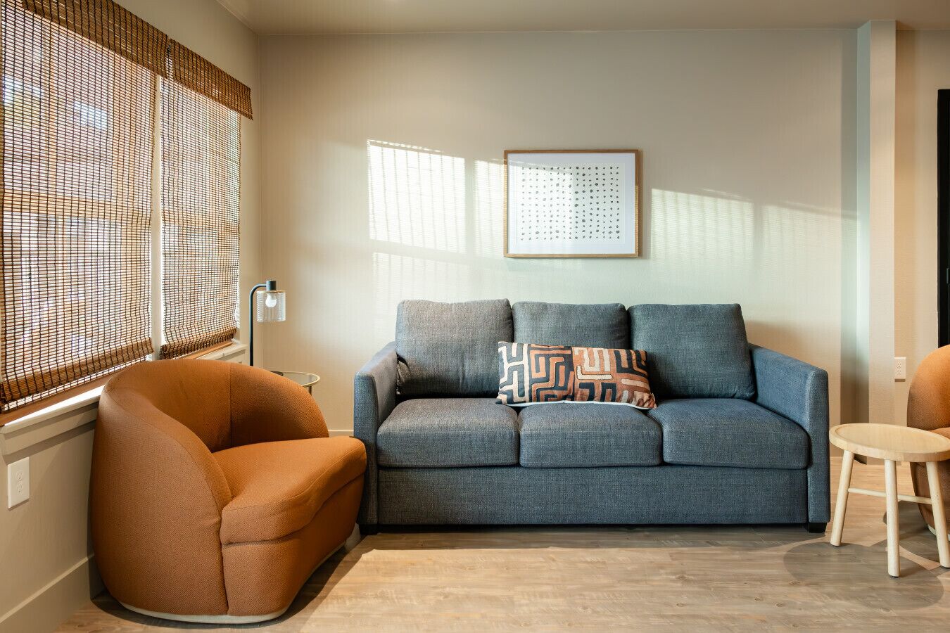 Een woonkamer met een grijze bank, een oranje fauteuil, een klein rond tafeltje en een raam met zonwering.