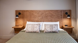 Habitación de hotel con cabecera de madera, dos mesitas de noche con lámparas, cama con almohadones estampados y colcha verde acolchada.