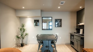 Un comedor con una mesa, sillas, una planta en maceta, un espejo y una zona de cocina con cafetera.