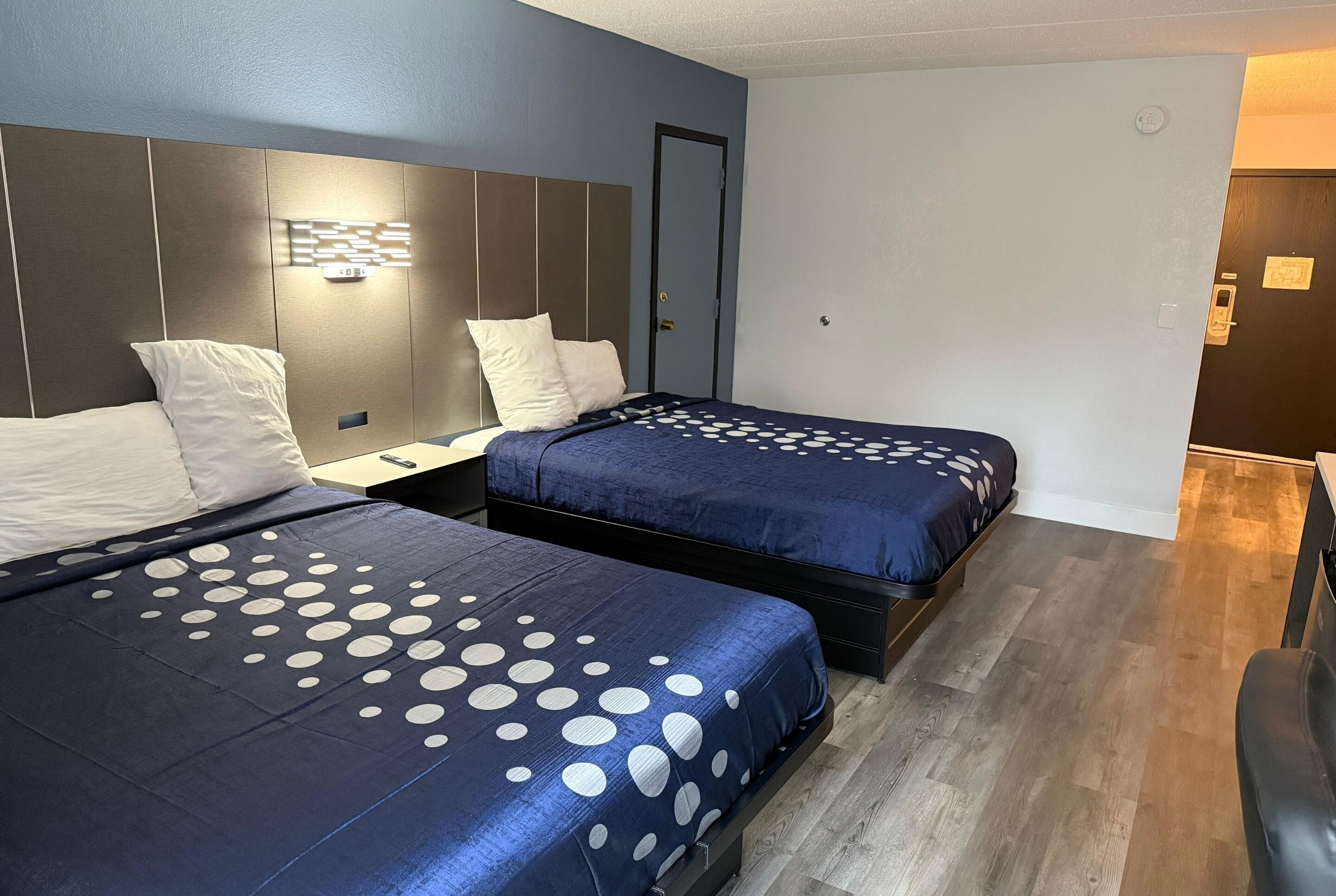 A hotel room with two beds, a nightstand, a wall-mounted light, and a door.