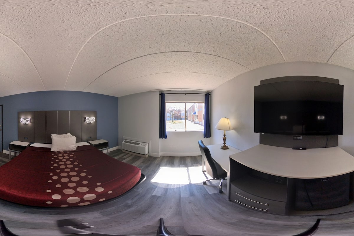 A room with a curved ceiling, a bed with a red bedspread, a TV, a desk, and a window with curtains.