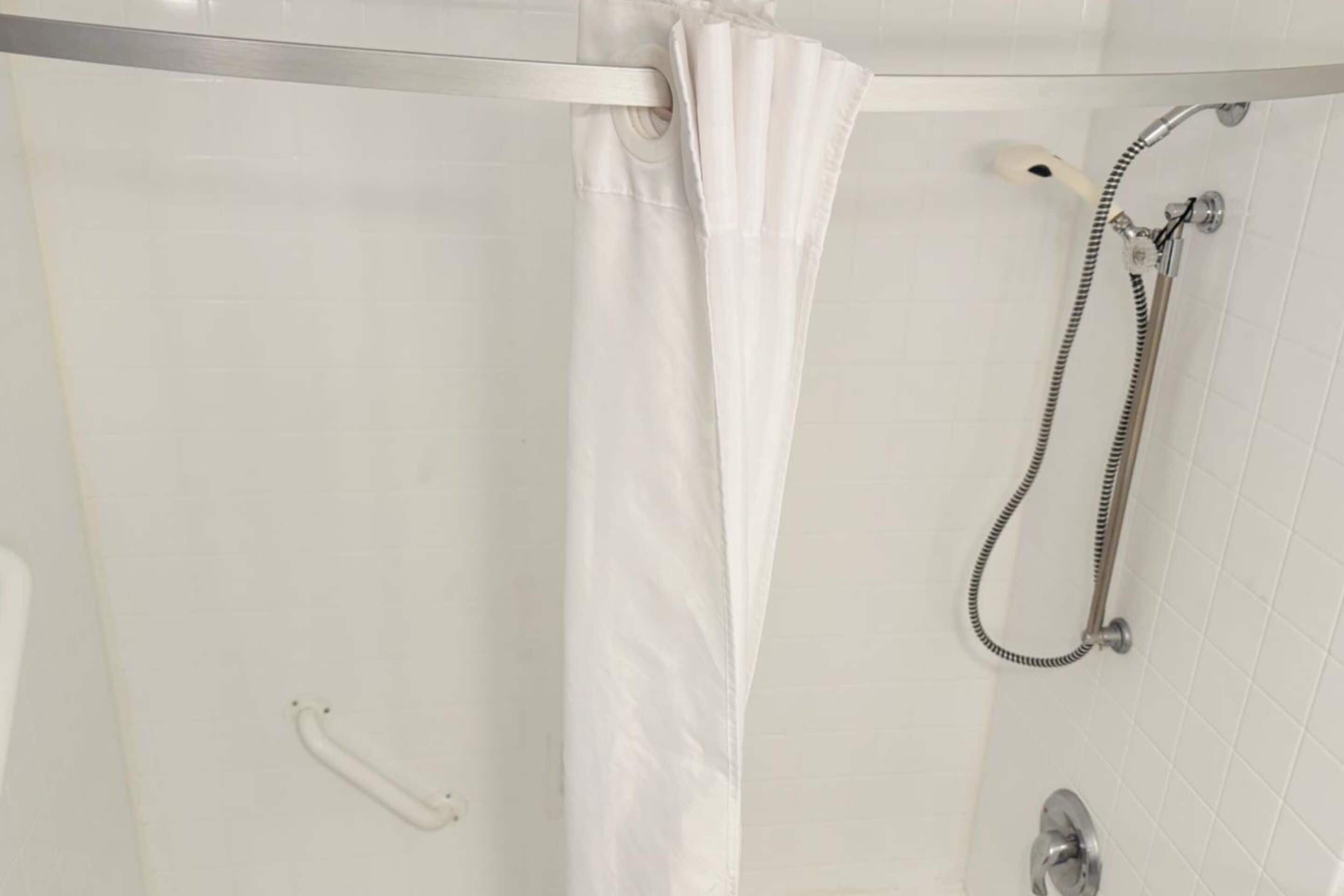 Un espace douche doté d’une porte en verre, d’un rideau de douche blanc, d’un pommeau de douche à main et d’un robinet mural.
