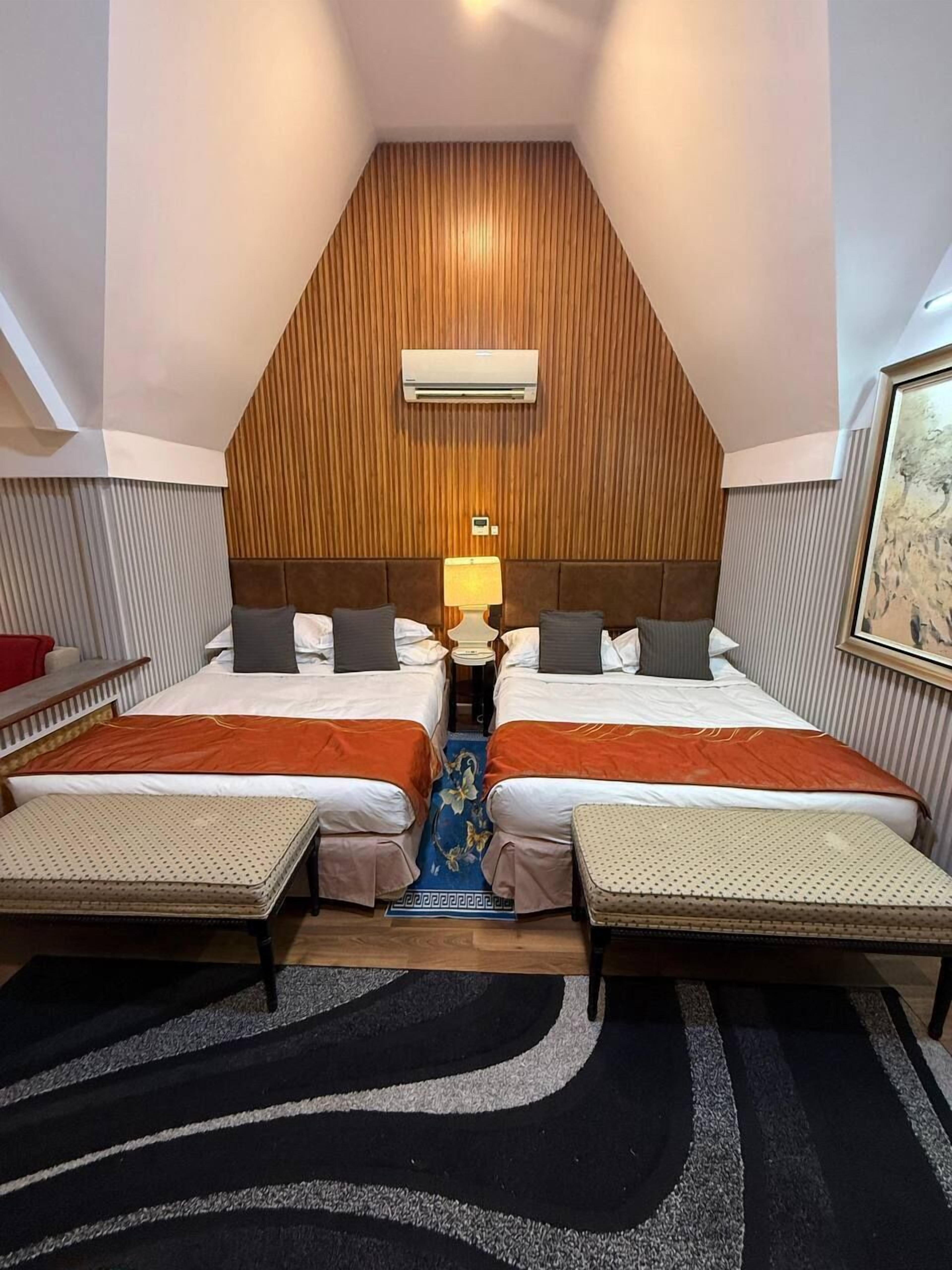 A hotel room with two beds, a wooden sloped ceiling, an air conditioner, a nightstand with a lamp, and a patterned rug.