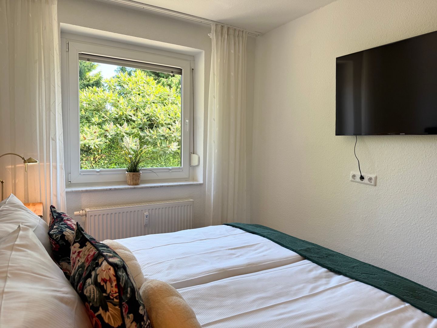 A bedroom with a bed, a window with curtains, a plant on the windowsill, a radiator, and a TV mounted on the wall.