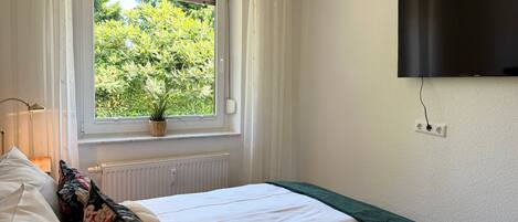 A bedroom with a bed, a window with curtains, a plant on the windowsill, a radiator, and a TV mounted on the wall.