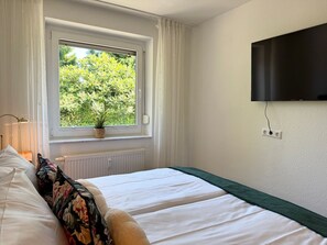 A bedroom with a bed, a window with curtains, a plant on the windowsill, a radiator, and a TV mounted on the wall. - Haus Strandgold - Strandgold 1 - Cozy vacation apartment with terrace and (Cuxhaven)