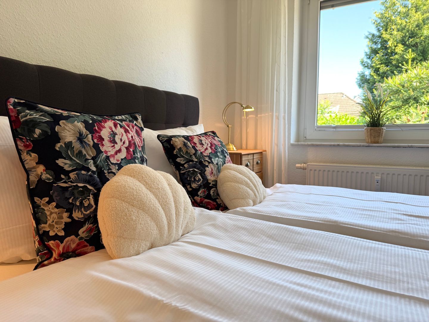 Ein ordentlich bezogenes Bett mit floralen Kissen und einem weißen Kopfteil in einem Zimmer mit Fenster und Heizkörper.
