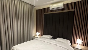 A modern bedroom with a large bed, two bedside tables with lamps, a ceiling-mounted air conditioner, and a window with curtains. - 106 AL-NAJEH JRE Apartments For Rent ave (Amman)