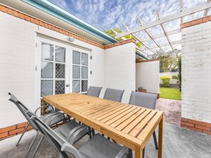 Un patio con una mesa y sillas de madera, un muro de ladrillo blanco y una puerta blanca con ventanal.