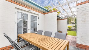 Un patio con una mesa y sillas de madera, un muro de ladrillo blanco y una puerta de cristal.