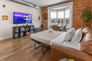 A modern living room with a flat-screen TV, a dartboard, a bookshelf with board games, a brown leather couch, and a window with a view of the outside.