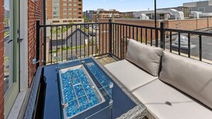 A balcony with a glass-top table, a cushioned seating area, and a view of surrounding buildings.