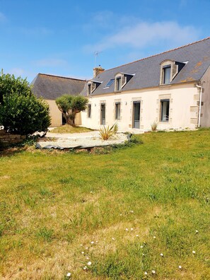 A two-story house with a sloped roof, a stone foundation, and a small garden area with grass and a tree. - LARGE SEASIDE FARMHOUSE FOR 10 PEOPLE (Tréogat)