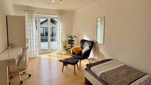 A modern bedroom with a bed, a desk, and a chair. There is a window with curtains, a plant, and a view of the outside.