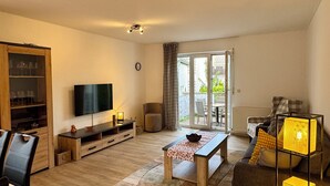 A spacious living room with a wooden floor, a dining table, a sofa, a TV, and a large window with a view of the outdoors.