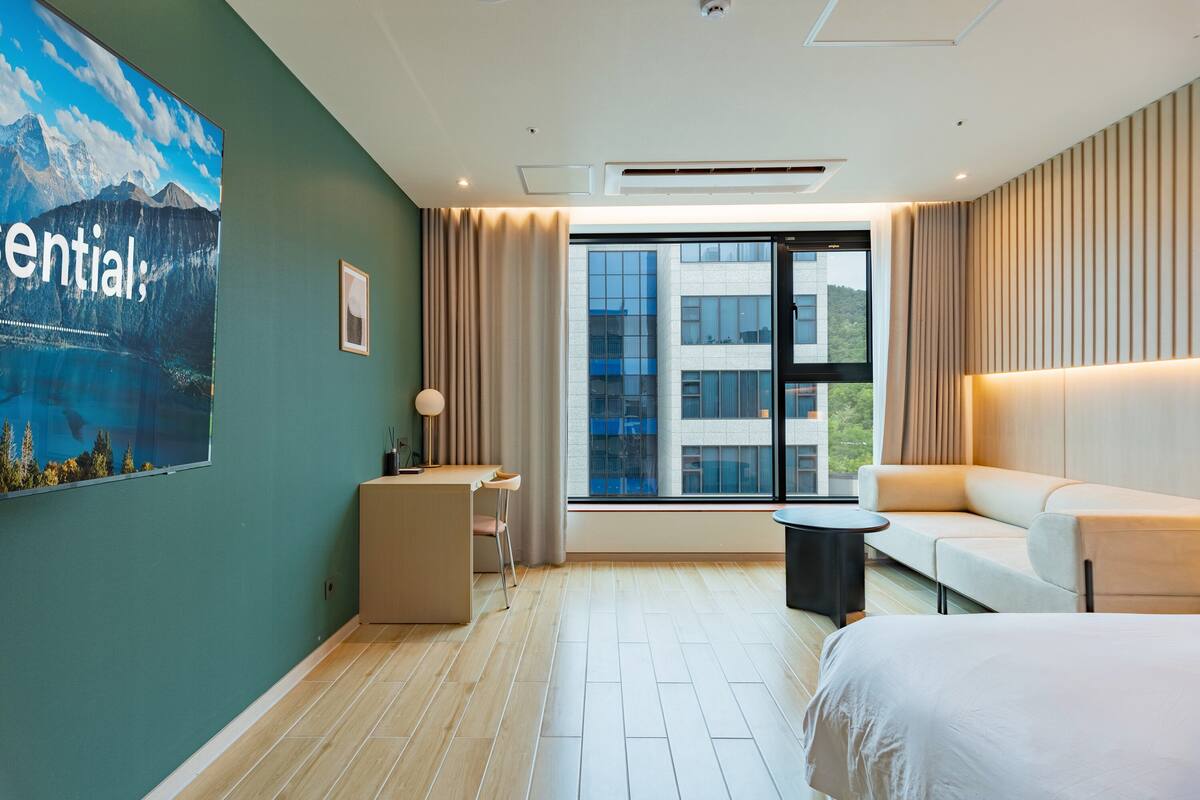 A modern hotel room with a large window, a bed, a sofa, a desk, and a chair.