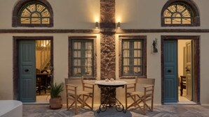Une terrasse extérieure avec une table ronde et quatre chaises, flanquée de deux portes dotées de fenêtres en arc de cercle, et un mur de pierre au centre.