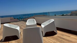 A rooftop terrace with white plastic chairs and a wooden floor, overlooking a coastal view.