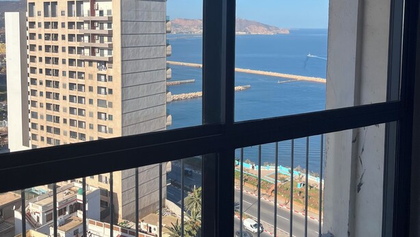 A view from a window overlooking a coastal area with a high-rise building, a road, and palm trees.