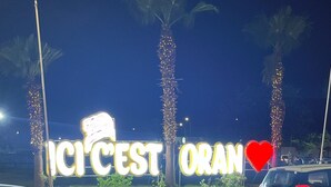 A parking lot with a sign reading "ICI CEST ORAN" and palm trees in the background.