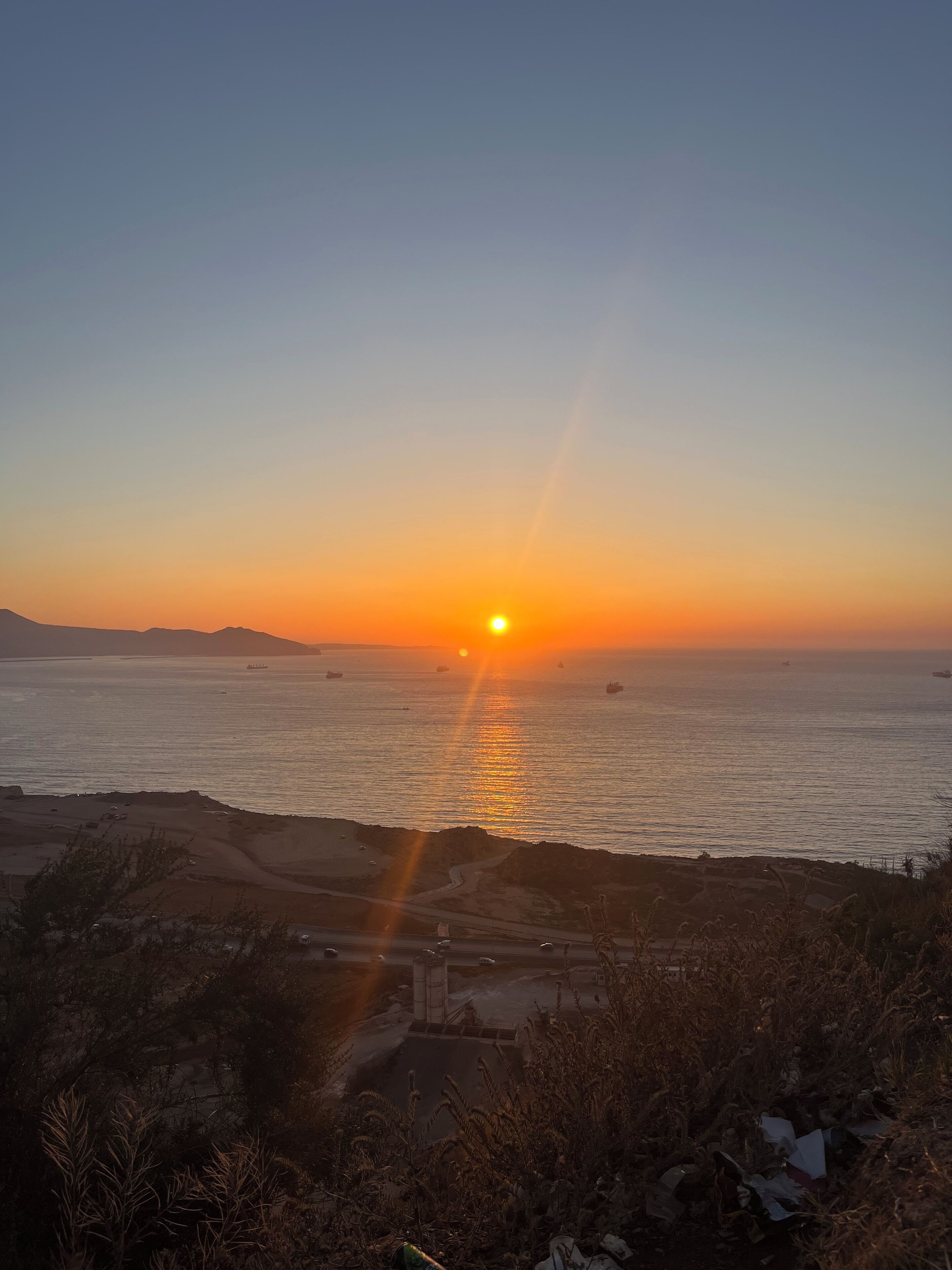 A sunset over a coastal area with a road and some vegetation in the foreground.