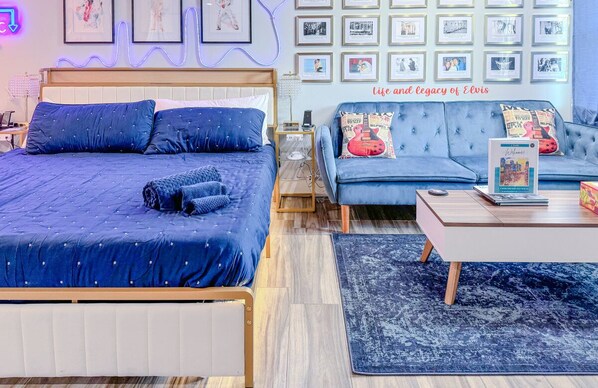 Un dormitorio con una cama, un sofá, una mesa de centro y una alfombra. Hay cuadros enmarcados en la pared y un letrero de neón.