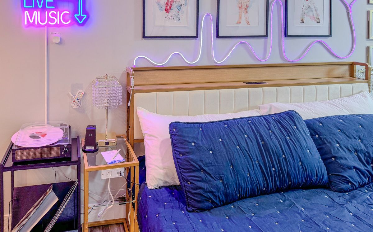 A bedroom with a bed, a bedside table with a lamp, a neon sign, and framed artwork on the wall.