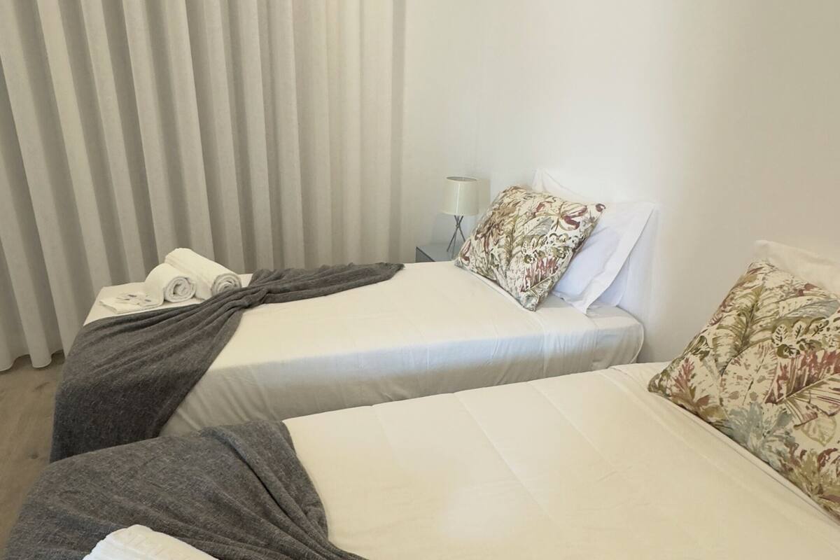 A hotel room with two beds, each with a white bedspread and a grey blanket. There are towels and a pillow on the beds.